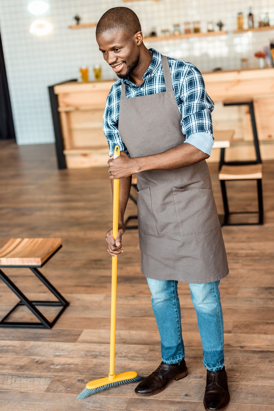 handsome smiling african american worker sweeping with broom in coffee shop