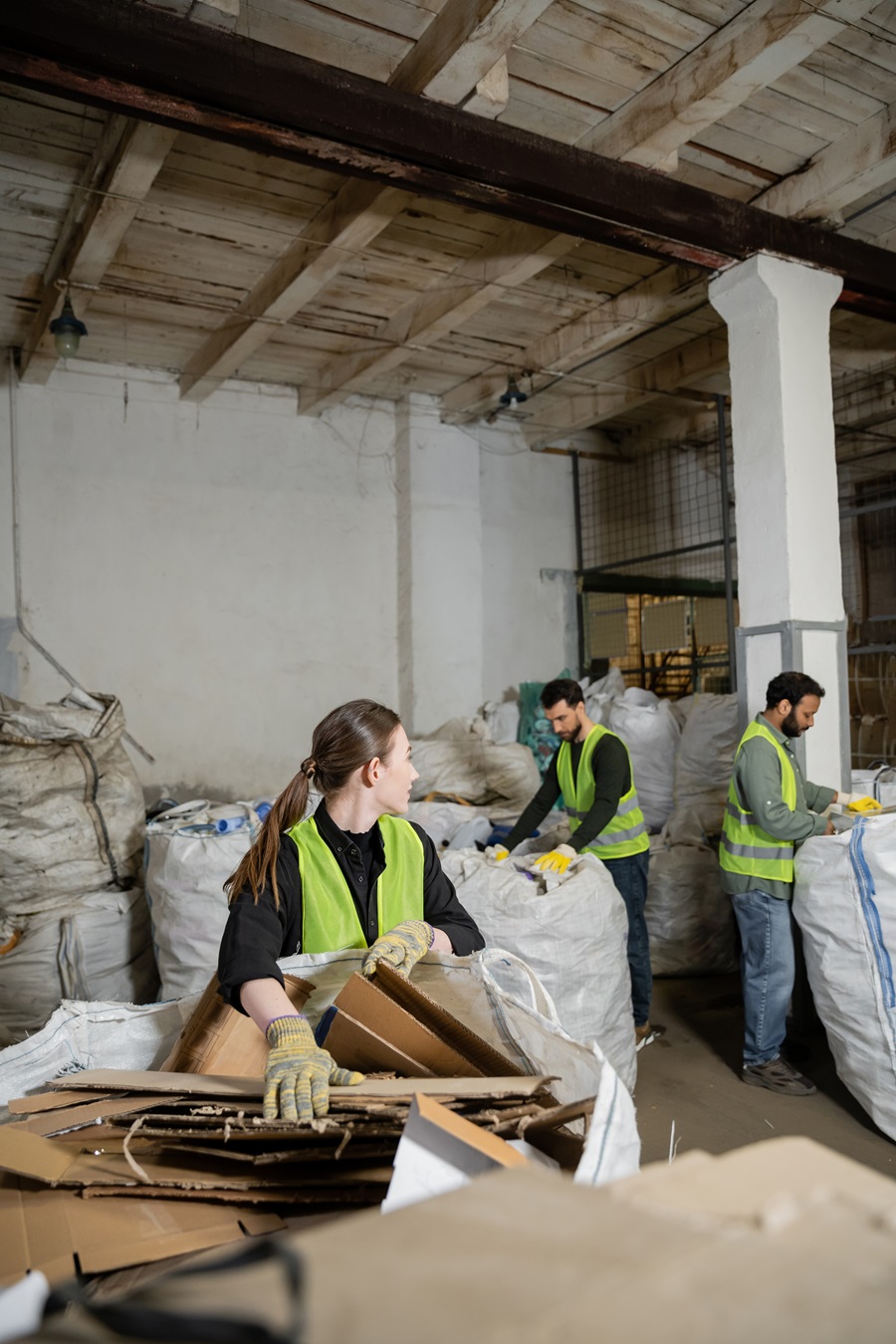Young female worker in protective gloves and vest looking at multiethnic colleagues while putting cardboard in sack in waste disposal station, garbage sorting and recycling concept